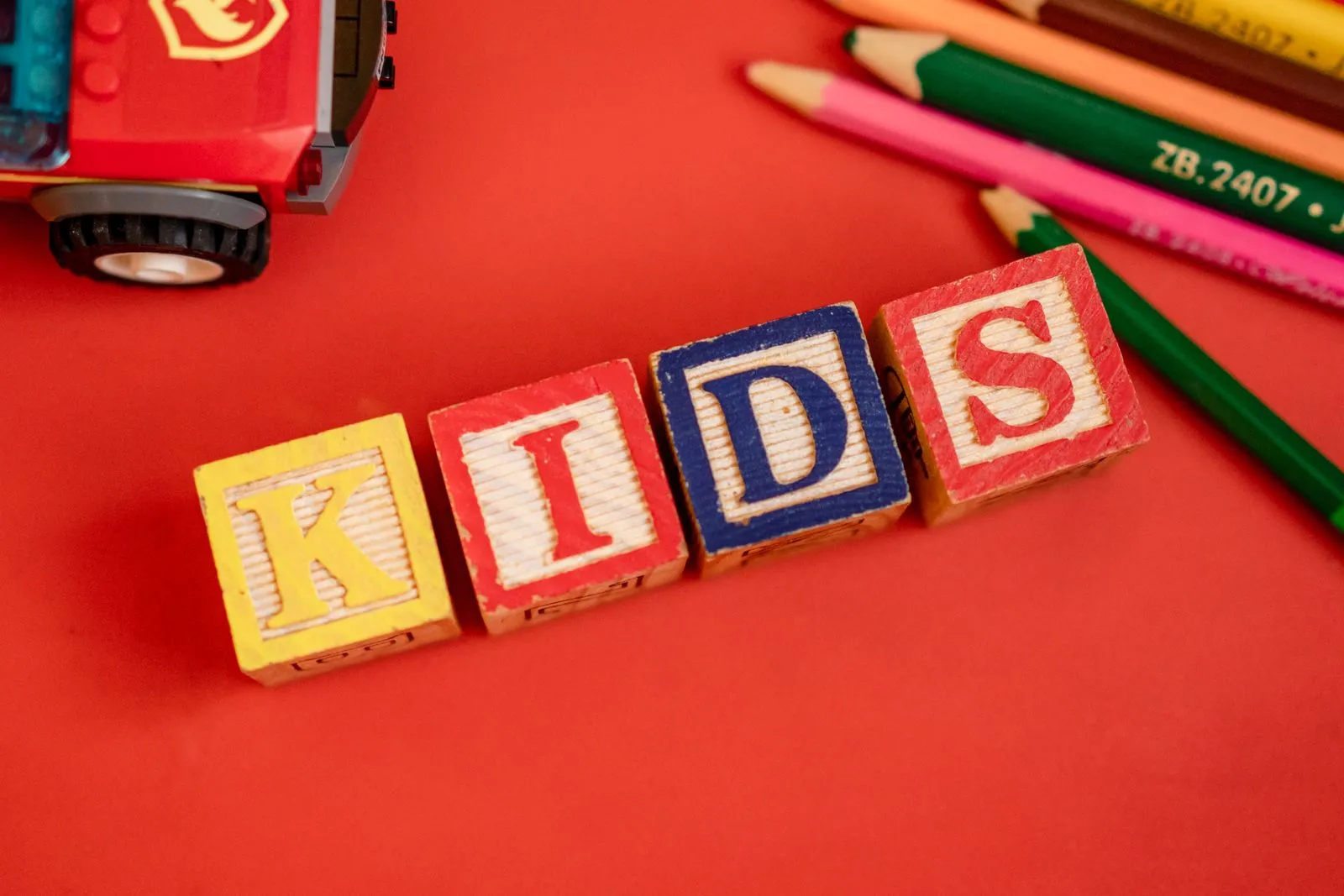 Creative layout of wooden blocks spelling 'KIDS' with pencils and toy on red background.
